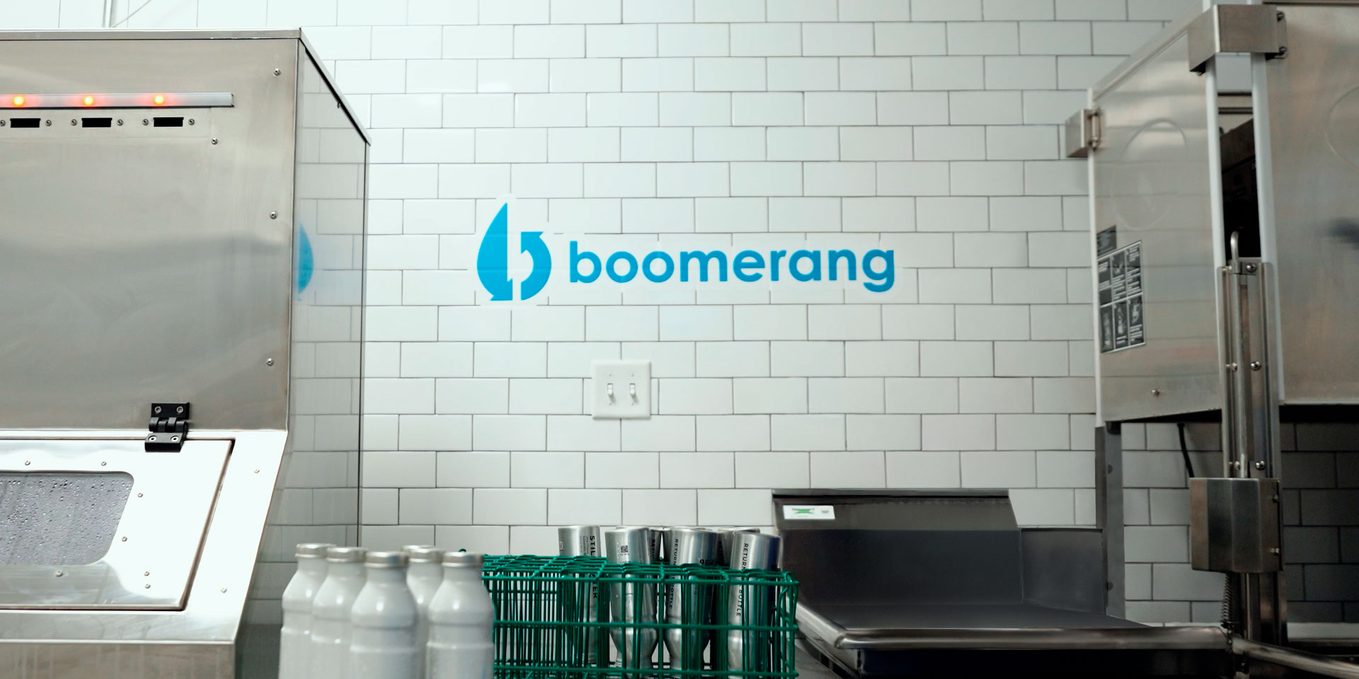 A commercial kitchen with white subway tile walls, stainless steel appliances, a green rack with metal cups, white bottles, and a blue "boomerang" logo with a leaf above it on the wall.