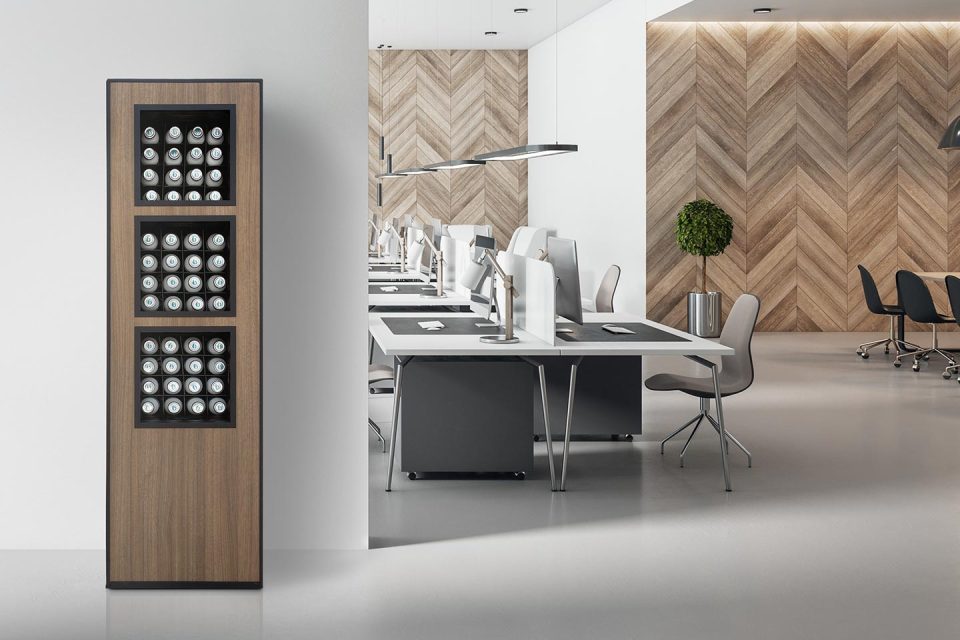 Modern office space with rows of desks and computers; in the foreground, a tall wooden cabinet with three glass sections displaying multiple beverage cans. Light, minimalist decor with herringbone wood wall and a potted plant.