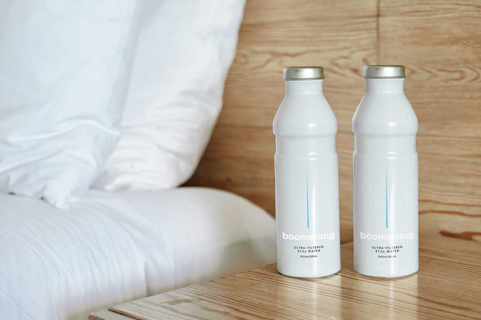 Two white reusable bottles labeled "boomerang ultra-purified still water" sit on a wooden nightstand next to a neatly made bed with white bedding and a wooden headboard.